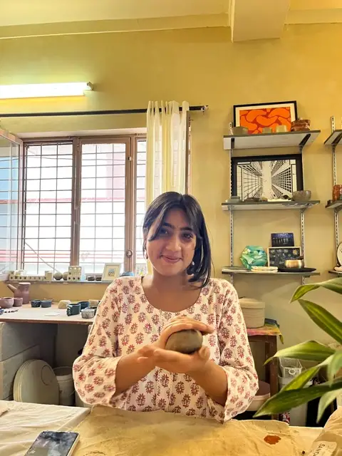 Harshita cradling a ball of clay in the sunlit studio