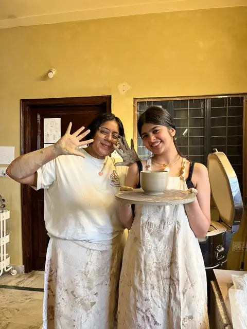 Two guests holding up the mug they threw, hands still covered in clay