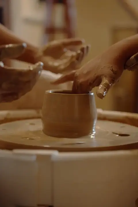 Two pairs of hands shaping a small cup together on the wheel