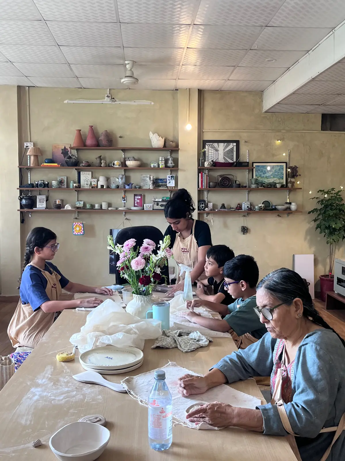 Harshita guiding a table of kids and elders through hand-building at the long studio table