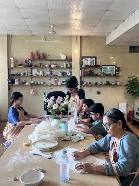 Harshita guiding a table of kids and elders through hand-building at the long studio table