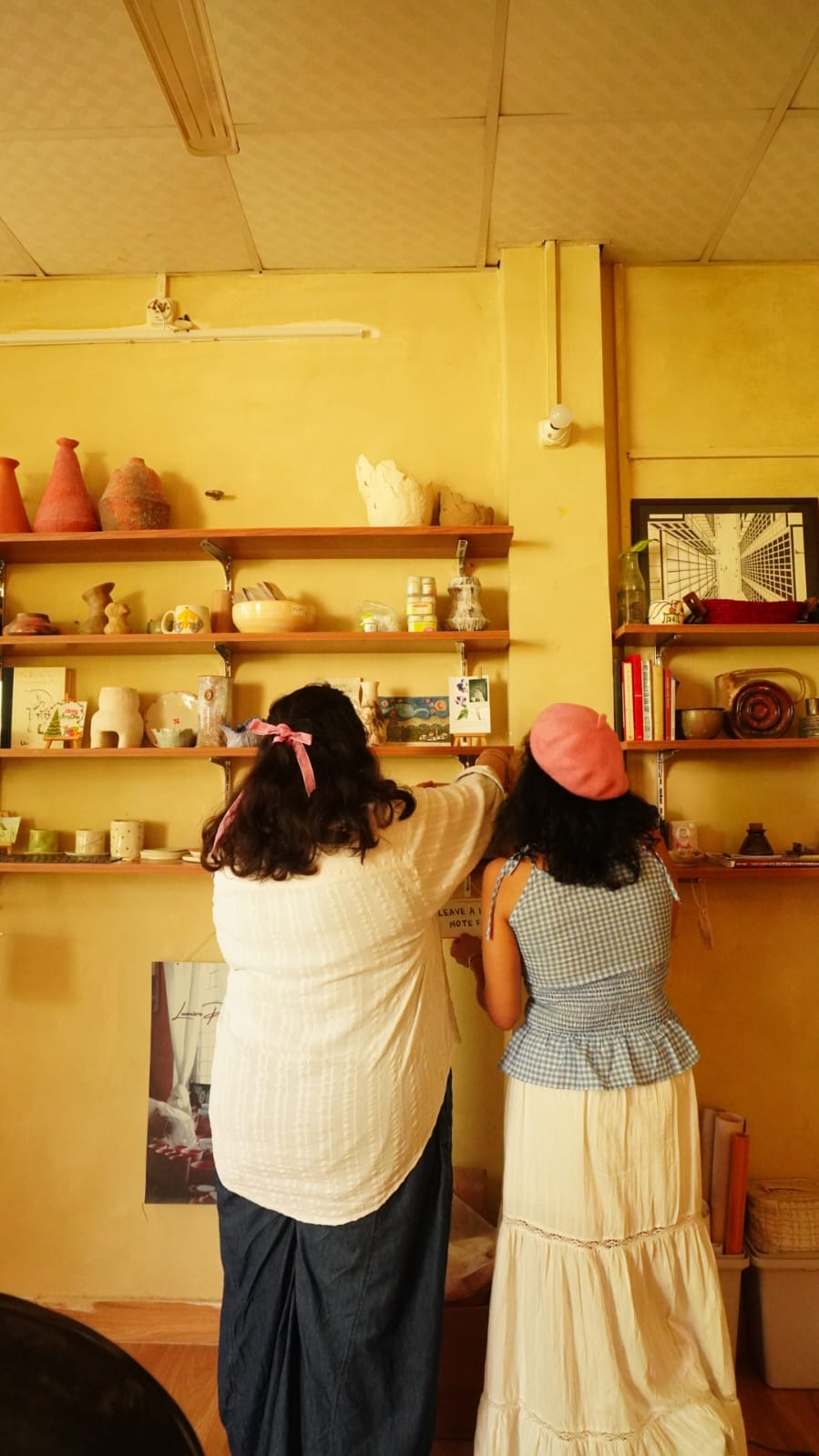 Two visitors pinning a note beside the studio's shelf of ceramics