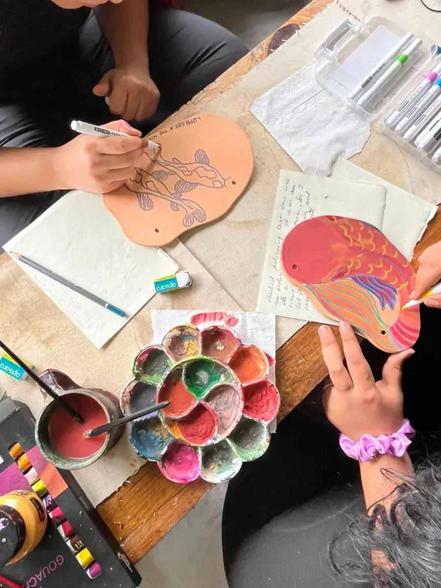 Hands painting fish motifs onto flat clay pieces at a wooden table