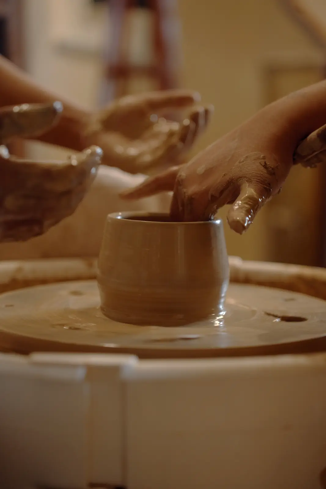 Two pairs of hands shaping a small cup together on the wheel