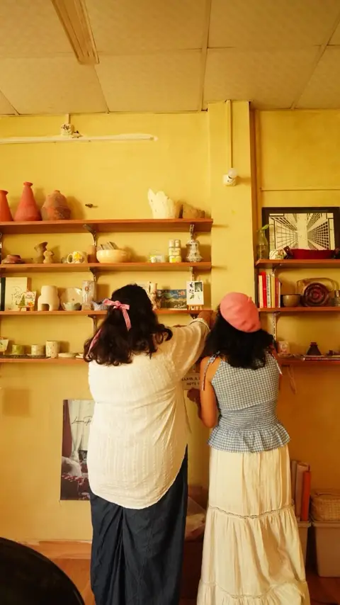 Two visitors pinning a note beside the studio's shelf of ceramics