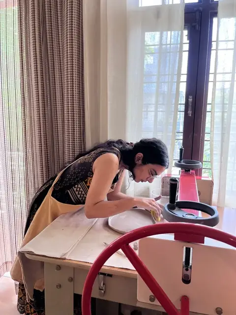 A maker leaning in to smooth a plate on the wheel by the window