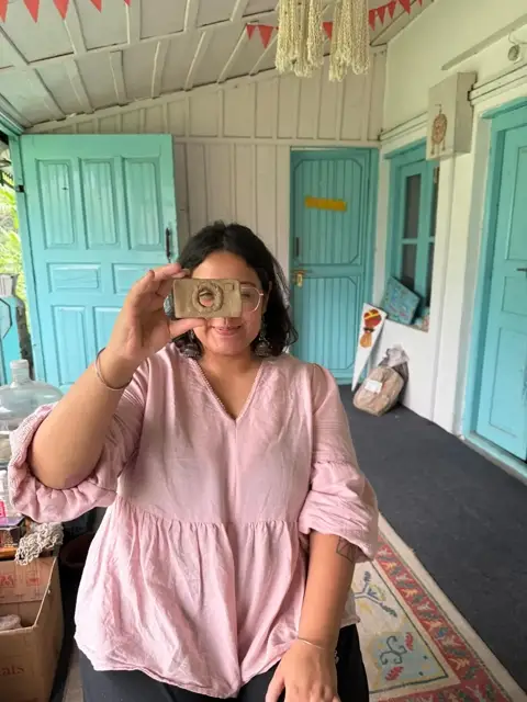 Harshita holding a clay viewfinder up to her eye on the studio verandah